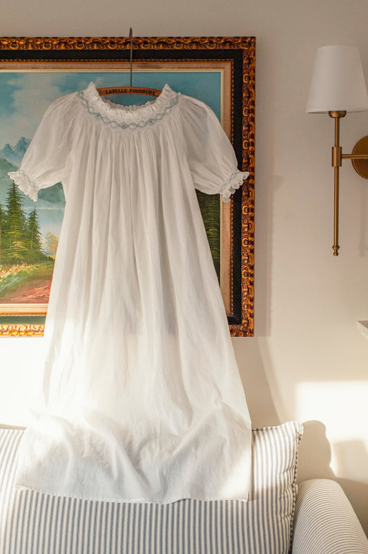 White hand-smocked heirloom girls nightgown with delicate blue embroidery, short puff sleeves, and a flowing 100% cotton voile silhouette, photographed hanging in warm natural light.